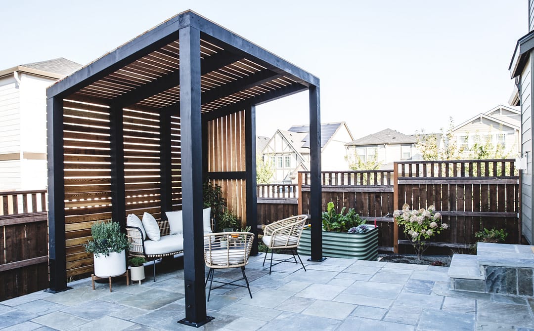 Minimalustic outdoor furniture under a metal framed pergola with wood slat walls and roof on a paver style patio