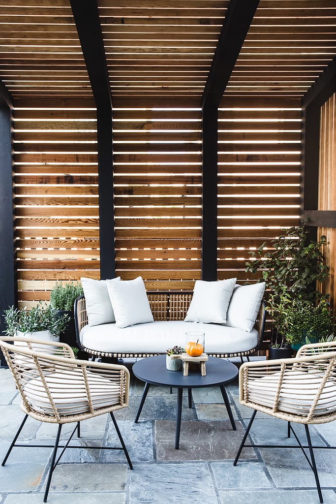 Minimalustic outdoor furniture under a metal framed pergola with wood slat walls and roof on a paver style patio