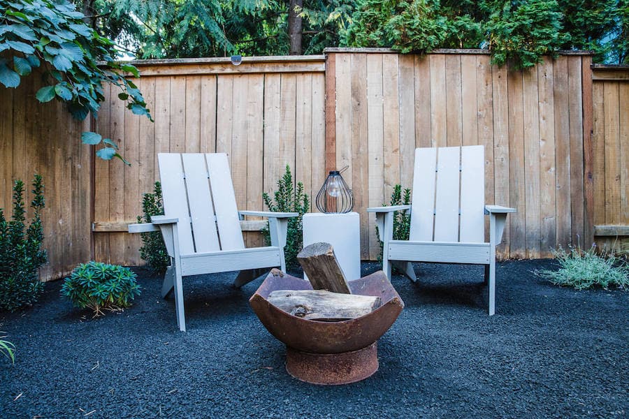 White adirondack chairs in the corner of a hardscaped yard with wooden privacy fence, minimal green plants, dark gray gravel and a small firepit